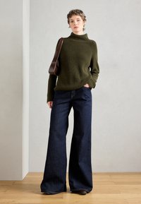 Un maglione a collo alto a coste color verde oliva abbinato a pantaloni a gamba larga blu scuro. Pavimento in legno naturale e parete neutra sullo sfondo.
