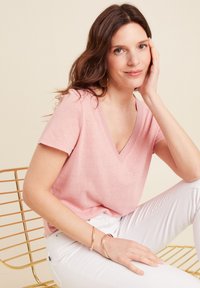 Rosafarbenes Baumwoll-V-Shirt mit V-Ausschnitt und kurzen Ärmeln, mit einem strukturierten Ausschnitt, kombiniert mit weißen Hosen. Accessoires sind dünne metallische Armbänder.