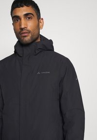 Giacca nera soft-shell con colletto alto, chiusura con zip e cappuccio. Presenta il logo Vaude sul petto e una texture liscia e opaca.