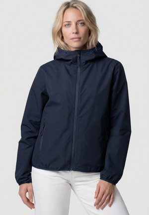 Blonde Frau trägt eine marineblaue Kapuzenjacke mit Reißverschluss und weiße Hose, steht vor einem einfarbigen hellen Hintergrund.