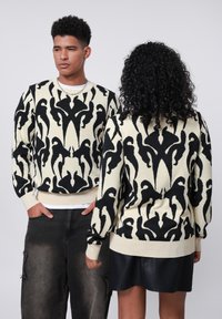 Strickpullover mit einem schwarzen abstrakten Muster auf einem cremefarbenen Hintergrund. Mit Rippbündchen an den Ärmeln und am Saum, lockere Passform. Kombiniert mit schwarzen Jeans und einem Rock.