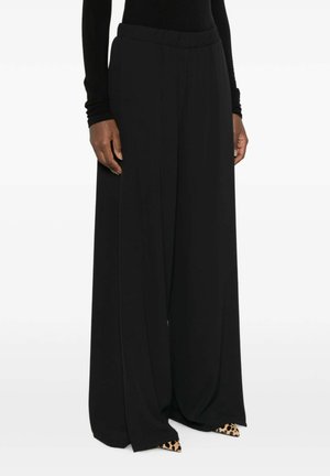 Donna che indossa pantaloni neri a gamba larga e top nero a maniche lunghe, con scarpe a punta a stampa leopardata visibili sotto i pantaloni.