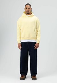 Lichtgele hoodie met een voorzak, een losse pasvorm en een capuchon met trekkoord, gedragen over een wit shirt. Donkere denim broek met wijde pijpen en laarzen.