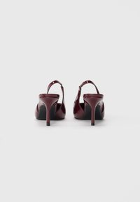 Escarpins en cuir verni bordeaux avec des talons hauts fins et un design à bout ouvert, présentant une découpe courbée au niveau du talon et une texture lisse.