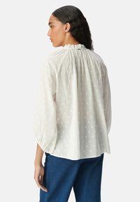 Camicia bianca con motivo a pois, tessuto leggero, collo arricciato e maniche a sbuffo, abbinata a jeans blu.
