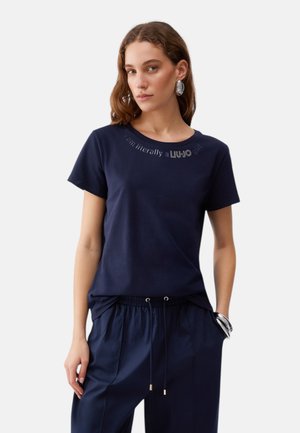 Donna con maglietta blu navy con testo in rhinestone vicino allo scollo e pantaloni blu navy abbinati con coulisse, accessoriata con orecchini e braccialetto d'argento.