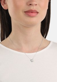 Silverhalsband med en blommig hänge med en liten ädelsten i mitten, bärs över en ribbad vit skjorta, vilande på en slät halslinje.