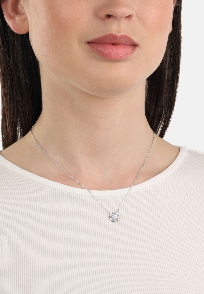 Silverhalsband med en blommig hänge med en liten ädelsten i mitten, bärs över en ribbad vit skjorta, vilande på en slät halslinje.