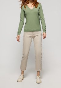Pull vert à col en V en matière douce, associé à un pantalon court de couleur crème. Le mannequin porte des baskets claires. Design simple, sans motifs visibles.