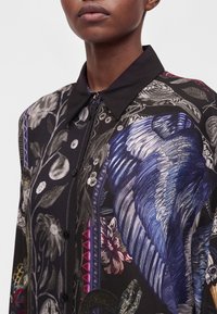 Gros plan d'une personne portant une chemise noire à col avec des motifs floraux et d'oiseaux détaillés en tons de bleu, gris et violet.