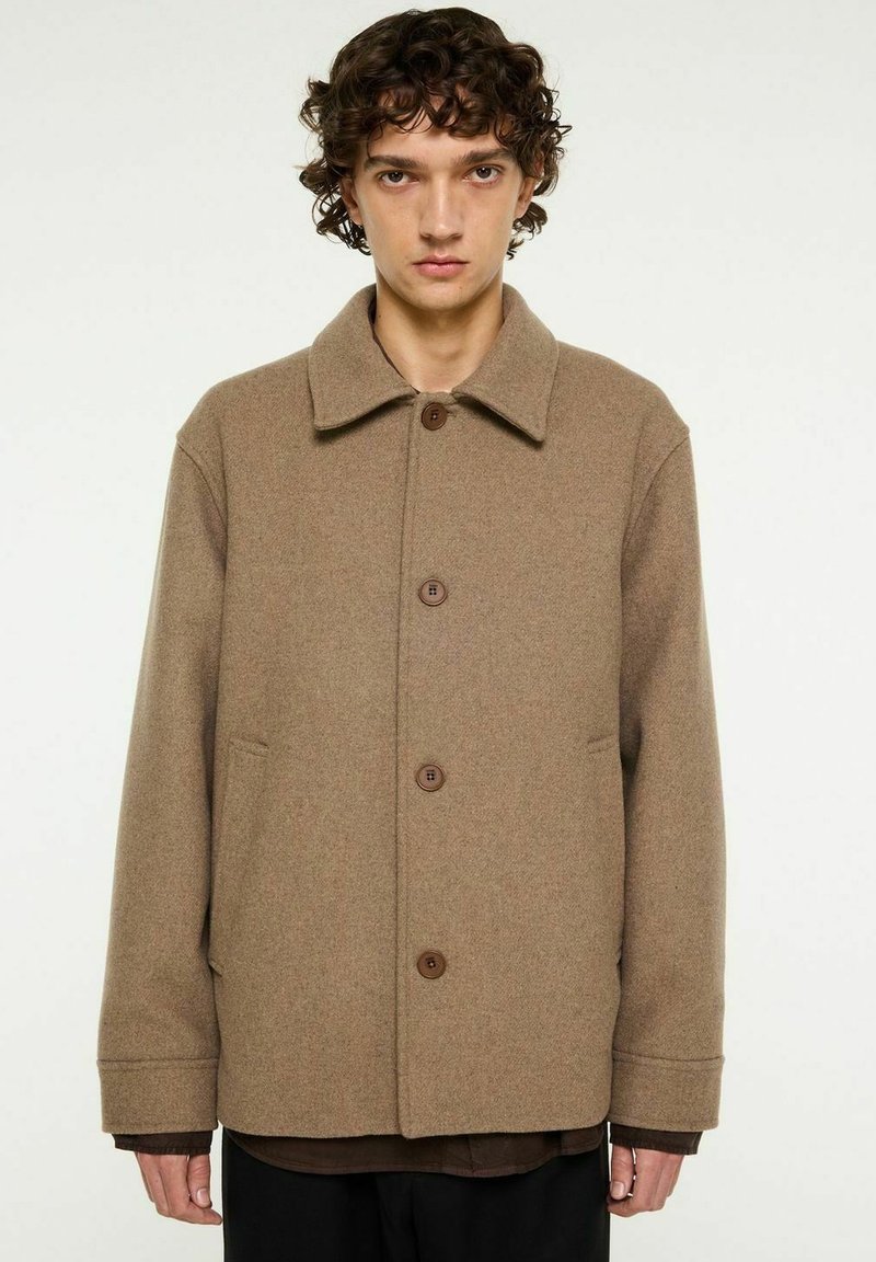 Brun ullkappa med rund krage, framknappstängning och sidofickor. Mjuk textur med avslappnad passform och minimal design.