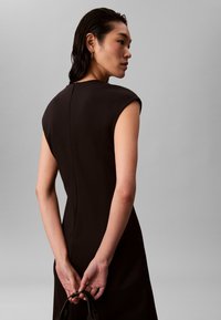 Svart figurnära klänning med ärmlösa cap sleeves, slät textur och en subtil söm bak. Modellen håller ett svart band i händerna och vänder ryggen mot kameran.