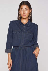 Camicia di jeans di colore blu scuro con colletto, tasche frontali e bottoni neri. Maniche lunghe arrotolate sui polsi.