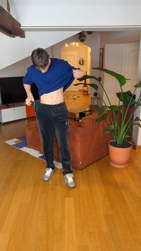 Νεαρός άνδρας σηκώνει μπλε πουκάμισο για να αποκαλύψει την κοιλιά του, προσαρμόζοντας μαύρο φόρμες σε σαλόνι με πορτοκαλί καναπέ, γλάστρα και ξύλινο πάτωμα.