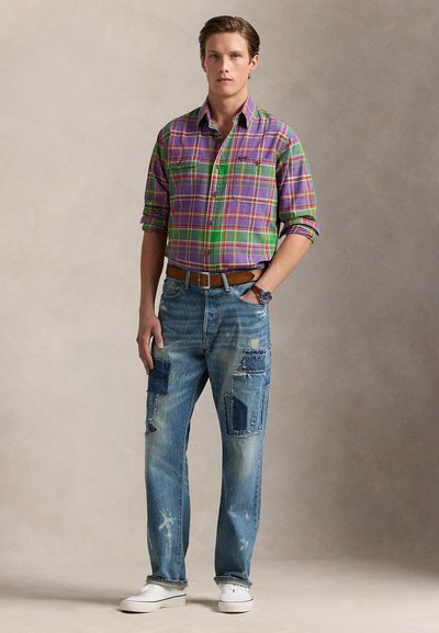 Tânăr stă în picioare purtând o cămașă în carouri mov și verde, blugi albaștri uzați cu petice, adidași albi și o curea maro, pe un fundal neutru.