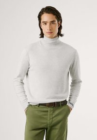 Maglione a collo alto grigio chiaro in morbido materiale knit, abbinato a pantaloni verdi e una cintura marrone con dettagli neri.