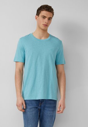 Junger Mann mit kurzen lockigen Haaren, der ein hellblaues T-Shirt mit kurzen Ärmeln und eine blaue Jeans trägt und vor einem schlichten grauen Hintergrund steht.