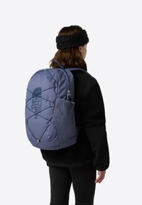 Lilla ryggsekk med teksturert overflate, med en logo og elastiske stropper. Den har en nettinglomme på siden og polstrede skulderstropper.