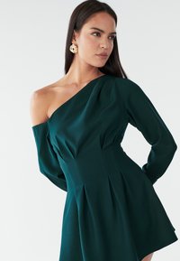Vestido verde de un solo hombro con mangas largas, falda plisada, tela suave, corsé ajustado y diseño drapeado. Se llevan pendientes de oro texturizados.
