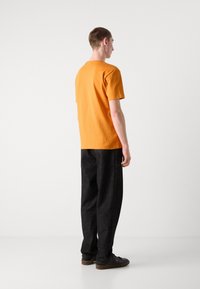 Orange bomulds T-shirt med klassisk rund hals, parret med sorte løse bukser, set fra ryggen, der viser et minimalistisk design.