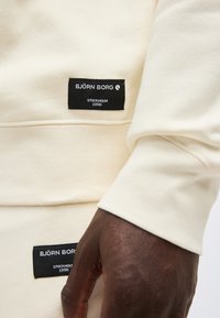 Χέρι με σκούρο δέρμα που ακουμπά κοντά σε μπεζ ένδυμα με μαύρες ετικέτες Björn Borg Stockholm 1956 στο μανίκι και στην περιοχή της μέσης.