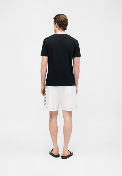 Άνδρας που στέκεται γυρισμένος από πίσω, φοράει μαύρο t-shirt, λευκά σορτς με κείμενο στο πλάι και μαύρα σανδάλια slide σε απλό λευκό φόντο.