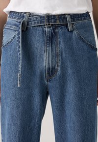 Denim jeans in light blue, featuring a standard fit with a visible belt, front pockets, and subtle stitching details. Textured fabric.