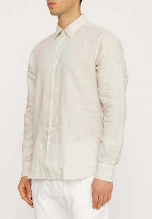 Camicia a maniche lunghe in lino beige chiaro con colletto button-down, caratterizzata da una trama sottile e un taglio dritto. Indossata con pantaloni bianchi.