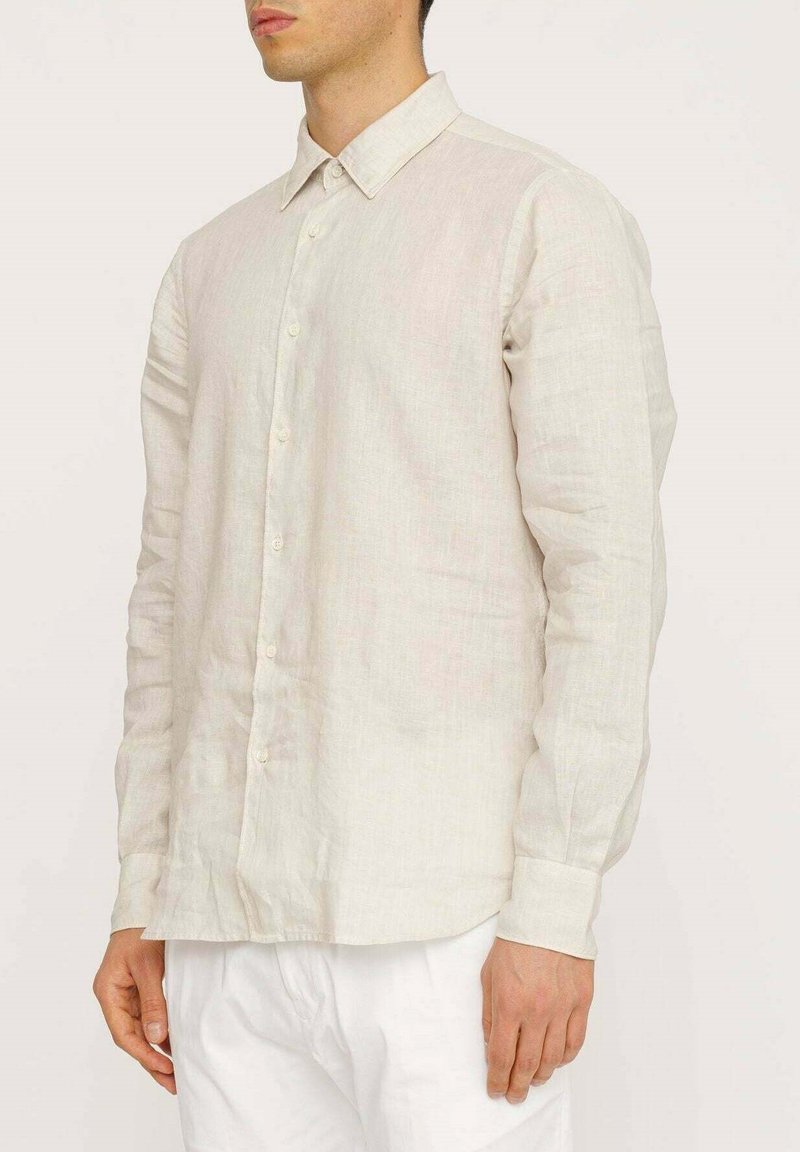 Camicia a maniche lunghe in lino beige chiaro con colletto button-down, caratterizzata da una trama sottile e un taglio dritto. Indossata con pantaloni bianchi.