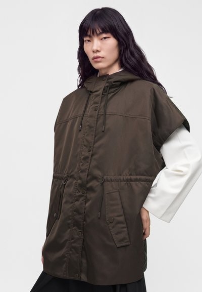 Jeune femme aux longs cheveux noirs portant une veste sans manches à capuche vert foncé par-dessus une chemise blanche à manches longues sur un fond uni.