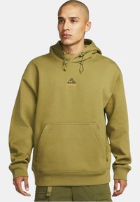 Olijfgroene hoodie gemaakt van zacht materiaal, met een buidelzak, trekkoorden en een klein logo in goud op de borst.