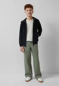 Czarna rozpinana bluza z szarym podszyciem, kremowa koszula z kieszonką, luźne zielone spodnie i białe sneakersy z brązowymi podeszwami.