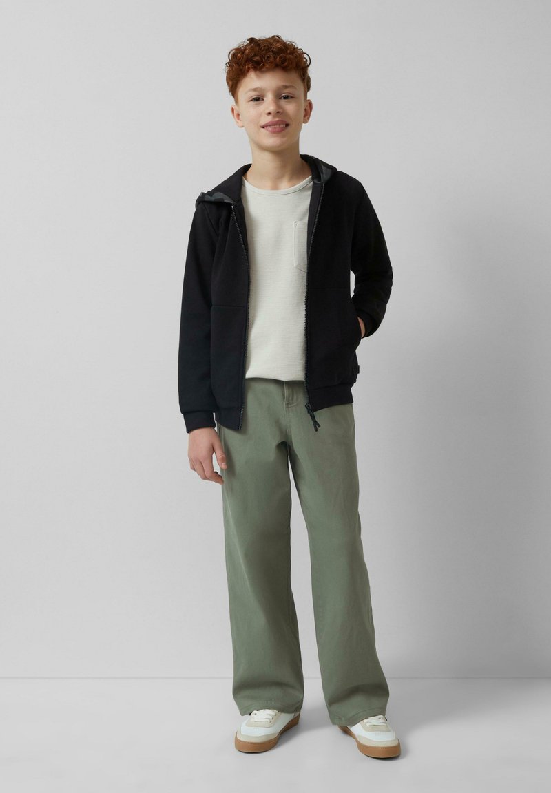 Czarna rozpinana bluza z szarym podszyciem, kremowa koszula z kieszonką, luźne zielone spodnie i białe sneakersy z brązowymi podeszwami.