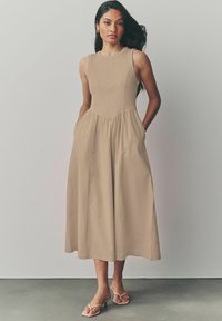 Beige ærmeløs kjole med tætsiddende overdel og flagrende skirt. Har sidelommer og ribbet tekstur øverst. Glat stof.