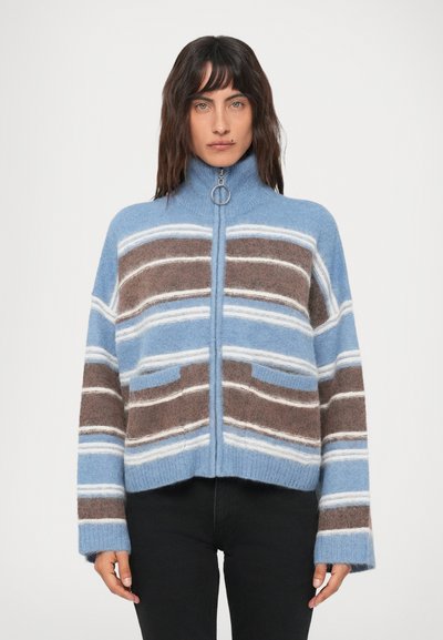 Woman wearing a blue, brown, and white striped zip-up sweater with a ring zipper pull and black pants, standing against a plain background.