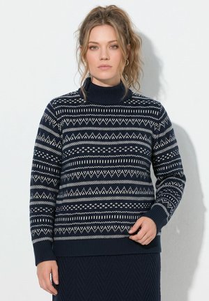 Kvinde med lyst bølget hår iført en marineblå og hvid mønstret højhalset sweater og mørkt skørt, stående mod en lyse baggrund.