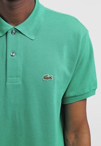 Grön polo-tröja i strukturerad tyg, med krage och två knappar. Litet, broderat krokodil-logotyp på vänster bröst.