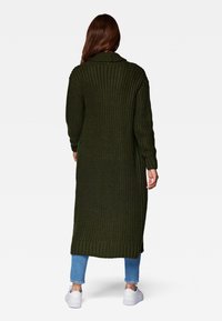 Lång, stickad kofta i mörkgrön med ribbat mönster, sjalkrage och sidofall, lagerpåsatt över blå jeans och vita sneakers.