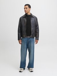 Mann steht und trägt eine schwarze Lederjacke über einem schwarzen Pullover und kariertem Hemd, weit geschnittene blaue Jeans und weiße Turnschuhe, schaut zur Seite.