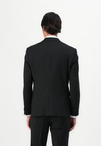 Veste de costume noire ajustée avec un design épuré, des épaules structurées, une fente arrière et des détails de poignet agrémentés de boutons.