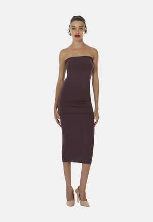Frau stehend, trägt ein trägerloses, dunkelviolettes, figurbetontes Midikleid und beige High Heels, mit zurückgebundenem Haar und rotem Lippenstift.