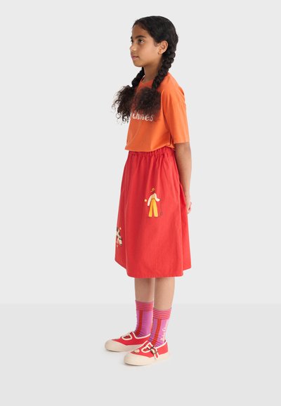 Fille aux cheveux tressés de profil portant un t-shirt orange, une jupe rouge avec des motifs, des chaussettes rayées violettes et des chaussures rouges.