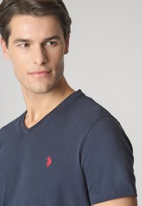 Maglietta a V blu navy in cotone, con un piccolo logo rosso sul petto. Il tessuto ha una superficie liscia e maniche corte.