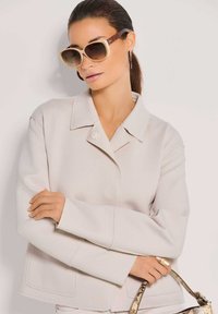 Frau mit einem eleganten Pferdeschwanz, die übergroße beigen Sonnenbrille, eine hellbeige Jacke, Kreolen und eine Handtasche mit Schlangenmuster trägt.