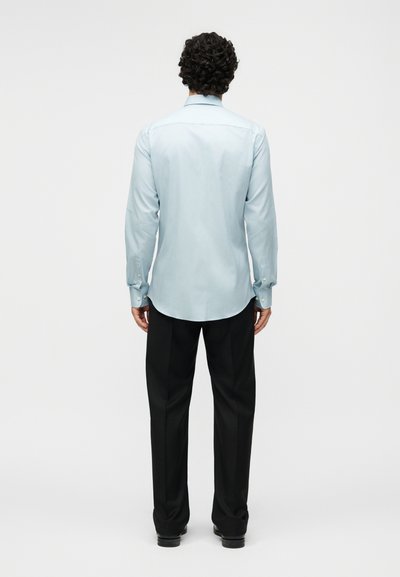 Άνδρας με σγουρά μαλλιά που φορά ένα ανοιχτό μπλε πουκάμισο με μακριά μανίκια και μαύρο παντελόνι, στέκεται με την πλάτη σε απλό φόντο.
