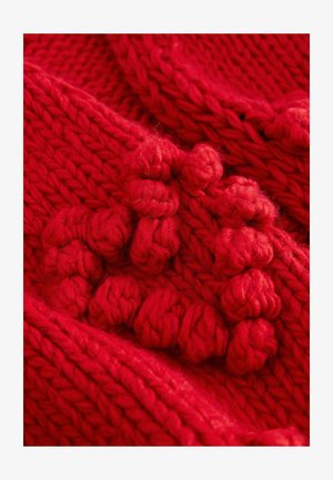 Rødt strikket tekstil med tykt garn og markante, hævede puf, der viser et struktureret og tæt mønster gennem hele stoffet.