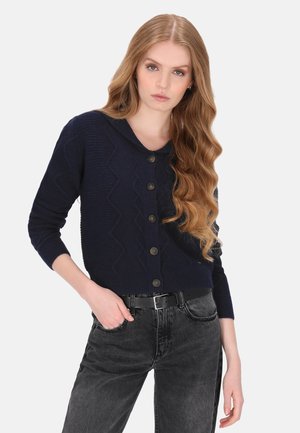 Kvinde med langt, bølget hår iført en mørk marineblå cardigan med knapper og sorte jeans med sort bælte, stående med den ene hånd i lommen.