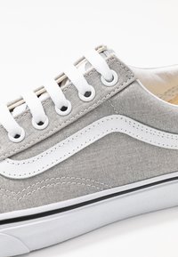 Šedé plátěné tenisky s bílými tkaničkami a bílým koženým vlnitým designem. Mají strukturovanou tkaninu a černou gumovou podrážku.