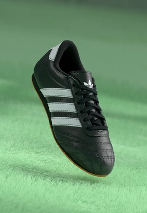 Baskets en cuir noir avec des rayures et des lacets blancs, semelle en gomme et logo Adidas sur la languette, présentées sur un fond texturé vert.