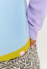 Sweter z kolorowym blokowaniem w jasnoniebieskim, lawendowym i zielonym kolorze. Posiada różową metkę z logo u dołu i jest zestawiony z dżinsami w panterkę.